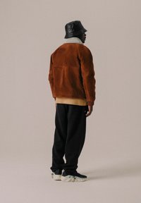 Chaqueta de ante marrón con cuello de borreguito crema, combinada con pantalones de chándal negros y un sombrero tipo bucket negro. El calzado presenta una suela clara.