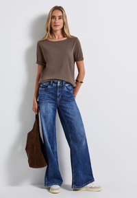 Bruine T-shirt met korte mouwen gecombineerd met wijde blauwe denim jeans. Het model houdt een donkerbruine tas vast; lichtgekleurde sneakers maken de outfit compleet.