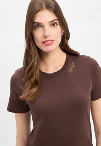 Bruine katoenen t-shirt met korte mouwen, met een ronde halslijn en een decoratieve slogan in rhinestones op de linkerschouder.