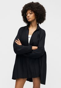 Junge Frau mit großen lockigen Haaren, die eine Brille, eine schwarze Jacke mit Reißverschluss und ein weißes Nike-Oberteil trägt, steht mit verschränkten Armen und schaut zur Seite.