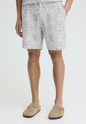 Personne portant un short gris clair à motifs avec des poches et des chaussures beige à enfiler, debout devant un fond uni clair.