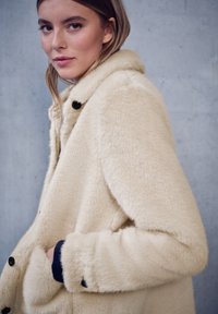 Manteau en fausse fourrure couleur crème avec un col, boutons noirs et surface texturée. Dispose de poches latérales et d'une coupe décontractée.
