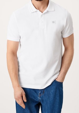 Mand iført en hvid kortærmet polo med en lille logo på brystet og blå denimjeans, den ene hånd i lommen.