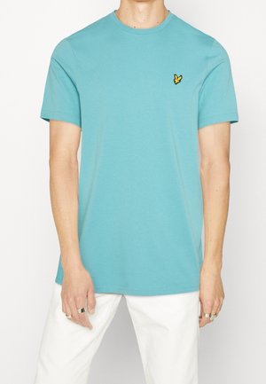 Mężczyzna ubrany w turkusowy t-shirt z krótkim rękawem z małym żółtym logo ptaka na piersi, w połączeniu z białymi spodniami i srebrnymi pierścieniami na palcach.