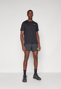 Mężczyzna stoi w czarnej koszulce Nike, czarnych szortach do biegania z żółtym napisem, czarnych skarpetach Nike i czarnych butach sportowych na szarym tle.