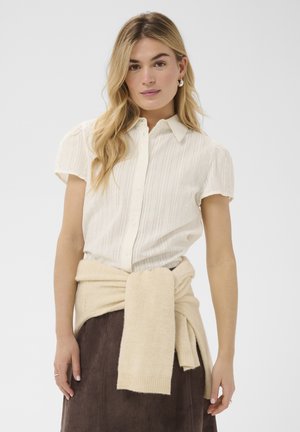 Jeune femme aux longs cheveux blonds portant une chemise blanche à manches courtes boutonnée, un pull beige noué autour de la taille et une jupe foncée.