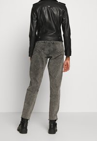 Personne portant une veste en cuir noire, un jean gris délavé, et des bottines noires se tenant contre un fond uni, de dos face au spectateur.