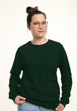 Junge Frau mit Brille und Hochsteckfrisur, trägt einen dunkelgrünen Pullover und hellblaue Jeans, hält den Pulloverbund, schaut nach rechts, neutraler Hintergrund.