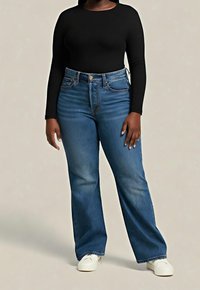 Personne portant un haut à manches longues noir, un jean bleu taille haute et des baskets blanches, debout devant un fond uni.