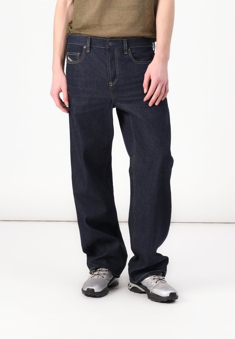 Personne portant un jean bleu foncé ample et des chaussures de sport argentées, debout devant un fond blanc.