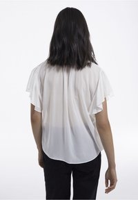 Vit, genomskinlig blus med lös passform, volangdetaljer på korta ärmar och rundad nederkant, i kombination med mörka byxor för kontrast.