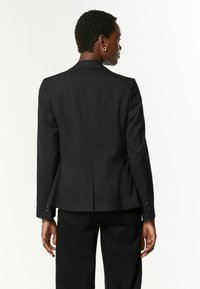 Černý blazer s přiléhavým střihem, s ostrými klopami, s jedním rozventilováním na zadní straně a knoflíkovými manžetami. Hladká textura látky.
