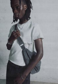 Hvid kortærmet t-shirt med broderet tekst, grå stof crossbody taske, mørk denim bukser, sølv kædetrøje og ringe.
