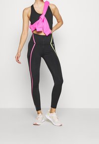Czarne legginsy z różowymi i neonowo-żółtymi paseczkami po bokach, w połączeniu z czarnym stanikiem sportowym i różową bluzą zawiązaną w talii. Białe trampki.