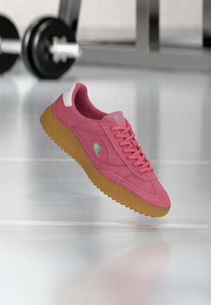 Pink suede sneaker with tan rubber sole and laces, featuring a small multicolor logo, set against a blurred gym background with weights.
