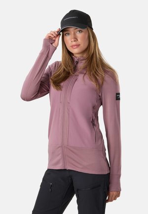 Femme ajustant une casquette noire, vêtue d'une veste mauve à fermeture éclair et d'un pantalon noir, sur un fond clair uni.