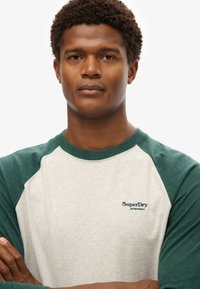 Szara i zielona koszulka raglanowa, z mieszanki bawełny, z okrągłym dekoltem i logo "SuperDry Athletics" na piersi. Widoczna teksturowana tkanina.