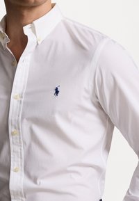 Wit overhemd met knopen en een klassieke kraag, voorzien van een klein blauw geborduurd logo op de linkerborst en een gladde katoenen textuur.