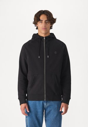 Zwarte zip-up hoodie gemaakt van zachte stof, met een trekkoord capuchon, voorzakken en een klein logo op de borst. Gepaard met blauwe jeans.