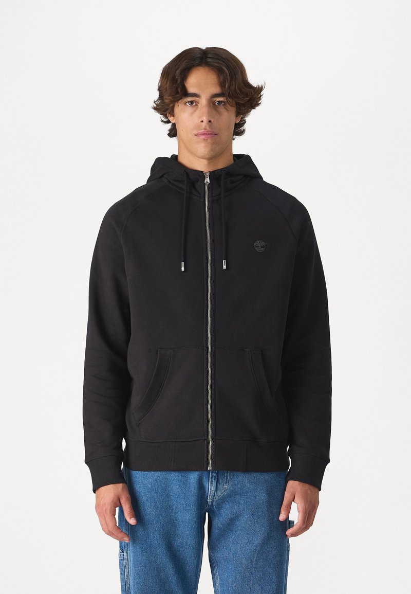 Felpa nera con zip realizzata in tessuto morbido, dotata di cappuccio con cordini, tasche frontali e un piccolo logo sul petto. Abbinata a jeans blu.