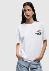 Camiseta blanca de algodón con un fit relajado, que presenta un bordado floral colorido en el lado izquierdo del pecho y el texto "Asociación China de Exposición Floral".