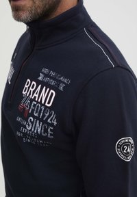 Tengerészeti félcipzáras fleece pulóver texturált anyagból, piros és fehér nyomott grafikákkal, valamint kör alakú logóval a ujján.