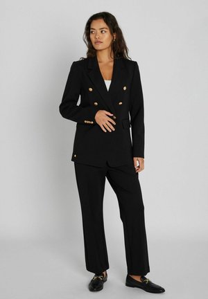 Femme debout portant un blazer noir croisé avec des boutons dorés, un pantalon noir assorti et des mocassins noirs, posant devant un fond uni.