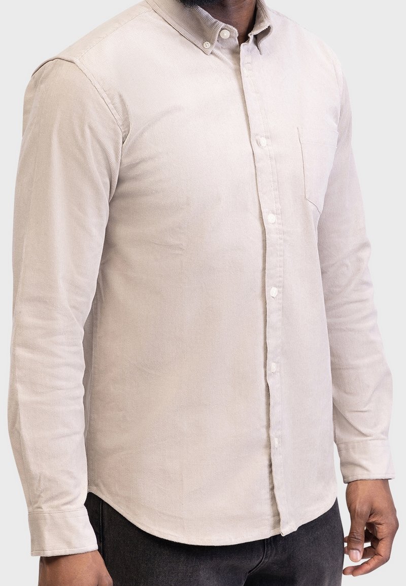 Homme portant une chemise beige en velours côtelé à manches longues avec des boutons et une poche, ainsi qu'un pantalon noir, debout devant un fond uni.