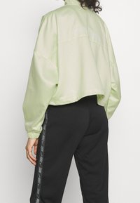 Ljusgrön kort kavaj i ett sidenliknande material med elastiska muddar, med en subtil logotypdetalj på baksidan. Kombinerad med svarta byxor.