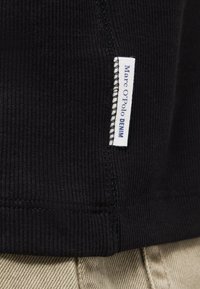 Μαύρο ριμπ ύφασμα με λευκή ραμμένη ετικέτα που γράφει "Marc O'Polo DENIM" δίπλα σε μπεζ υφή υφασμάτινη επιφάνεια.