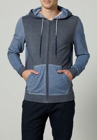 Pánská mikina na zip s kapucí, která kombinuje tmavé a světlé odstíny modré barvy, s texturovanou tkaninou, předními kapsami a stahovací šňůrkou na kapuci.