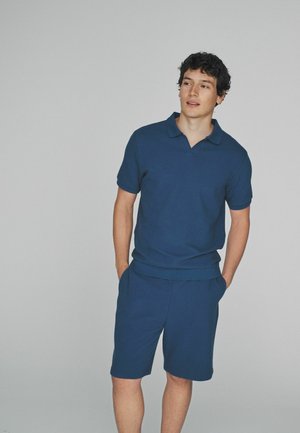 Fiatal férfi zsebre tett kézzel, kék rövid ujjú galléros pólót és rövidnadrágot viselve, kissé oldalra nézve.