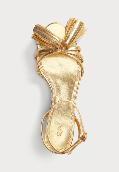 Sandale en cuir doré avec une semelle plate, des brides entrecroisées et un gland décoratif sur le devant. Fini métallique lisse avec un détail de marque.