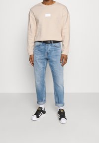 Beige sweatshirt with a square white label, light blue denim jeans, rolled cuffs, and black-and-white sneakers with gold accents.