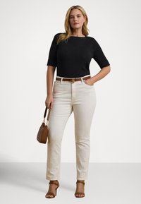 Μαύρο εφαρμοστό πουλόβερ, εκρού τζιν, καφέ ζώνη και τσάντα χειρός. Τα σανδάλια με ψηλό τακούνι ολοκληρώνουν την εμφάνιση. Το μοντέλο στέκεται με αυτοπεποίθηση, τα χέρια στις τσέπες.