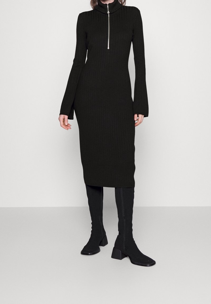Femme portant une robe midi noire moulante à côtes avec une fermeture éclair devant et des manches longues, associée à des bottes noires montantes à talons.
