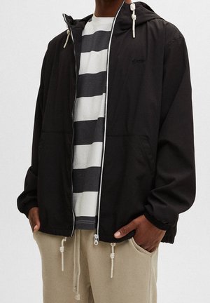 Persona con sudadera negra con cremallera sobre camiseta a rayas gris y blanca con las manos en los bolsillos de los pantalones beige, de pie frente a un fondo liso.