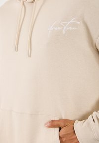 Beżowa bluza z kapturem, z kieszenią z przodu i sznurkami, z białym, kursywą napisem "Twój ruch" na piersi oraz ręką częściowo włożoną do kieszeni.