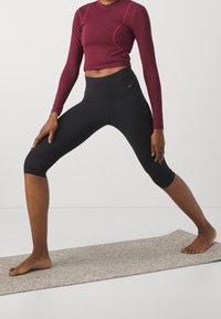 Top lungo bordeaux a maniche lunghe con cuciture rosa, abbinato a leggings neri a vita alta. Il modello è in piedi su un tappeto testurizzato a piedi nudi.
