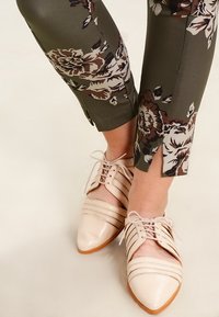 Světle béžové kožené oxford boty s špičatými špičkami a dekorativními výřezy, doplněné tmavě zelenými kalhotami s květinovými vzory.