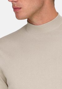 Suéter de alto cuello color beige claro, hecho de tela acanalada, con una silueta ajustada y textura suave. Primer plano del área del cuello.