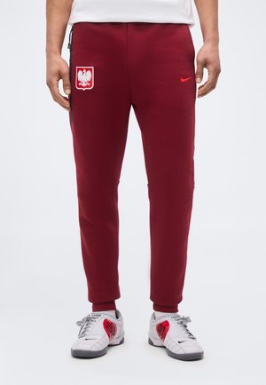 Pantalon de jogging bordeaux avec écusson aux armoiries de la Pologne et logo Nike rouge porté avec des baskets Nike blanches et rouges sur une personne debout.