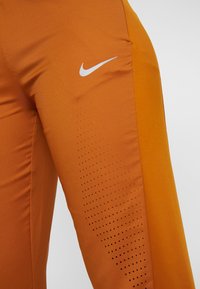 Orangefarbene Sporthose aus dehnbarem Stoff, mit einem weißen Nike-Logo, Panelakzenten und einem strukturierten, perforierten Design an der Seite.
