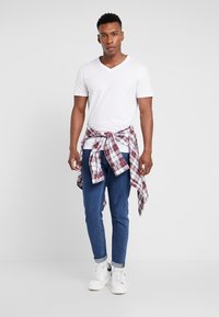 Hvid v-hals t-shirt, blå denim jeans, rød- og hvidternet flannel skjorte bundet om taljen og hvide sneakers. Enkel design og afslappet stil.