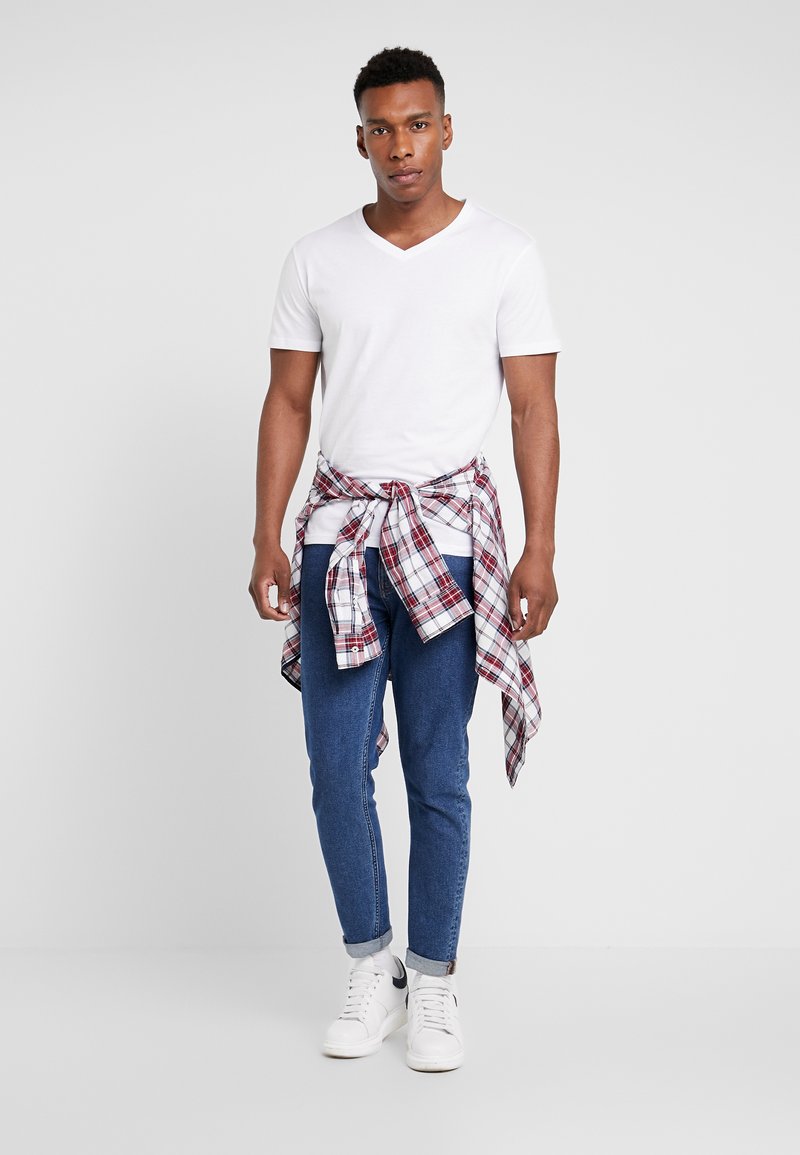 Hvid v-hals t-shirt, blå denim jeans, rød- og hvidternet flannel skjorte bundet om taljen og hvide sneakers. Enkel design og afslappet stil.