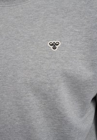 Grauer Stoff mit einem kleinen, eingestickten schwarz-weißen Bienenlogo, das mittig auf dem Brustbereich eines Kleidungsstücks positioniert ist.