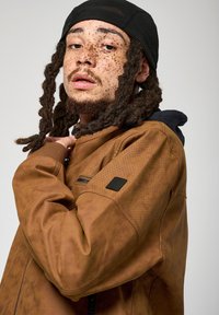 Veste perforée marron avec capuche, dotée d'une poche zippée et d'un patch logo noir sur l'épaule. Le tissu a une finition texturée.