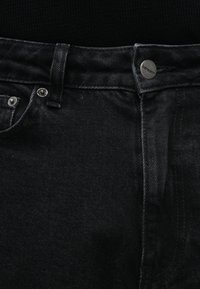 Jeans de ganga preta com uma ligeira textura, botões metálicos e um cós cosido. O tecido tem um brilho subtil e durabilidade.