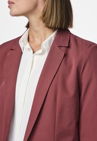 Femme aux cheveux courts et raides portant une chemise blanche boutonnée et un blazer mauve, tournée vers la gauche sur un fond blanc uni.