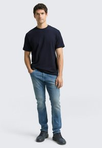 Granatowy t-shirt z krótkim rękawem zestawiony z jasnoniebieskimi obcisłymi dżinsami i czarnymi sznurowanymi butami. Model stoi na neutralnym tle.